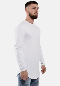 Wit T-shirt met lange mouwen en een ronde hals, gemaakt van lichtgewicht stof. Heeft een gebogen zoom en duimgaten bij de mouwen.
