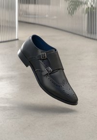 Scarpa elegante in pelle nera con doppia fibbia, punta affusolata, dettagli perforati e silhouette slanciata. Texture liscia e design moderno.