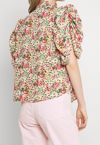 Blouse fleurie avec des manches ballon ; fond rose avec des fleurs rouges et vertes ; tissu doux ; ourlet arrondi ; associée à un pantalon rose clair.