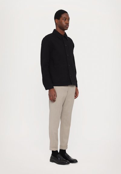 Black cotton jacket with button closure and two front pockets, paired with light beige cuffed trousers and black shoes.