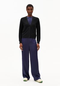 Cardigan nero a maglia con scollatura a V, sei bottoni, indossato sopra una tuta satinata blu navy. Pantaloni larghi con una texture liscia. Sneakers bianche e verdi.