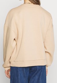 Sudadera beige de gran tamaño con cuello redondo, costuras de hombros caídos, puños acanalados y un ajuste relajado, combinada con jeans de mezclilla oscura.