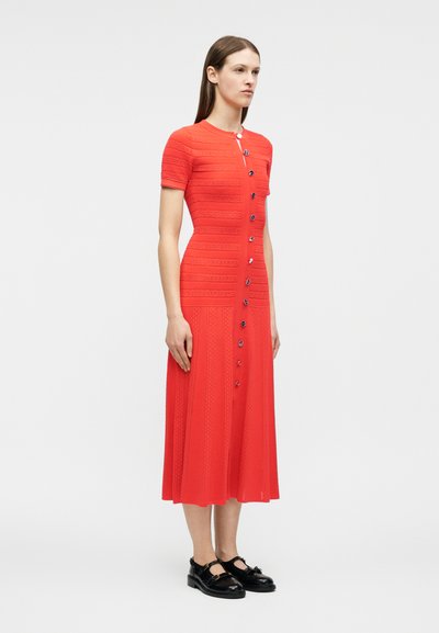 Γυναίκα που φοράει εφαρμοστό, κόκκινο φόρεμα με κοντά μανίκια και μήκος μέχρι τον αστράγαλο, με κουμπιά στο μπροστινό μέρος και μαύρα παπούτσια Mary Jane, στέκεται μπροστά σε απλό φόντο.