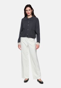 Dunkelgrauer, verkürzter Cardigan mit spitzem Kragen, Knöpfen vorne und zwei Vordertaschen, kombiniert mit weißen, weit geschnittenen Hosen und schwarzen Ballerinas.