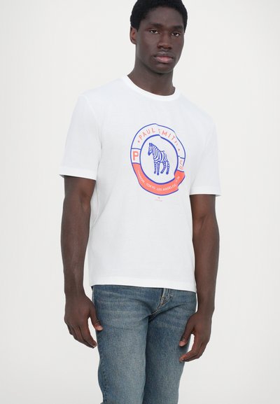 T-shirt en coton blanc avec un imprimé graphique d'un zèbre dans un logo circulaire, des accents bleus et oranges, et des manches courtes. Porté avec un jean bleu.