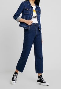 Mörk denimjacka med uppkavlade ärmar, knäppning framtill, kombinerad med högmidjade mörka jeans och svarta högklackade sneakers. Vit grafisk t-shirt synlig under.