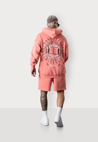 Haut à capuche rose avec un design de soleil et du texte dans le dos, associé à un short assorti et des sneakers blanches. Avec une jambe tatouée et des lunettes de soleil.