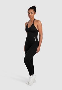 Schwarzer athletischer Bodysuit mit gerafftem Detail an der Brust, kombiniert mit schwarzen Leggings. Weiße Logoakzente an der Seite und am Knöchel.