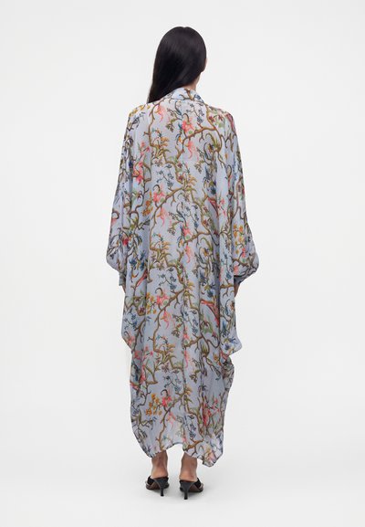 Femme aux longs cheveux foncés portant une robe ample longue au sol avec un motif coloré de fleurs et de branches, associée à des sandales noires à talons.