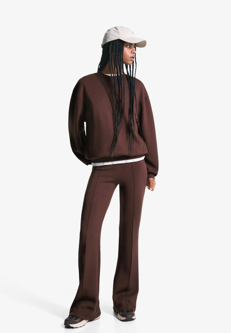 Bruine sweatshirt en bijpassende flared broek van zacht materiaal; voorzien van witte stikselaccenten. Draagt met een lichte pet en sneakers.