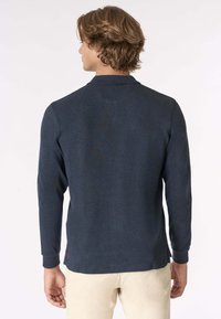 Polo a maniche lunghe di colore navy con tessuto texturizzato, orlo leggermente arrotondato e un logo discreto vicino alla cucitura laterale inferiore.