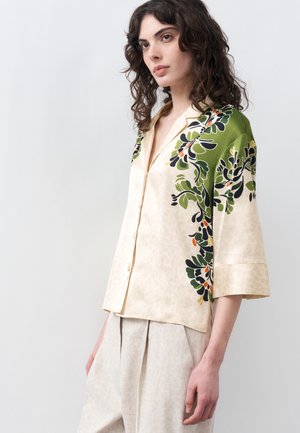 Νεαρή γυναίκα που φοράει κρεμ μπλούζα με κουμπιά, με πράσινο και μαύρο floral σχέδιο στους ώμους και ανοιχτό μπεζ παντελόνι.