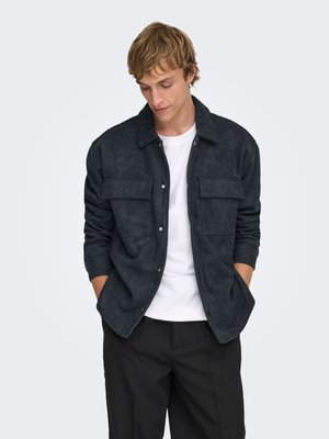 Giacca in suede grigio scuro con collo, chiusura con bottoni e due tasche sul petto. Indossata sopra una camicia bianca e abbinata a pantaloni neutri.