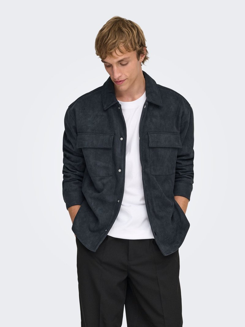 Veste en daim gris foncé avec un col, une fermeture par boutons et deux poches poitrine. Portée par-dessus une chemise blanche et associée à un pantalon de couleur neutre.