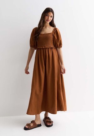 Bruine linnen maxi-jurk met een smock-bodice en pofmouwen. Gecombineerd met bruine sandalen met een geweven design.