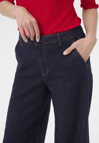 Tummansiniset farkut, joissa on korkea vyötärö, viisi taskua ja keltaiset tikkausleikinnät. Kangas on sileäpintaista.