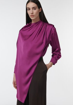 Mujer vistiendo una blusa asimétrica de satén magenta con hombro abotonado, combinada con pantalones negros de tiro alto, posando frente a un fondo liso.