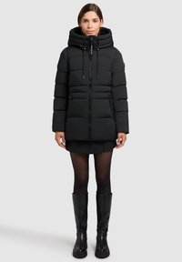 Sort puffjakke med hætte, der har lynlås foran og to sidelommer. Materialet ser blødt ud med et quiltet design og en elegant overflade.