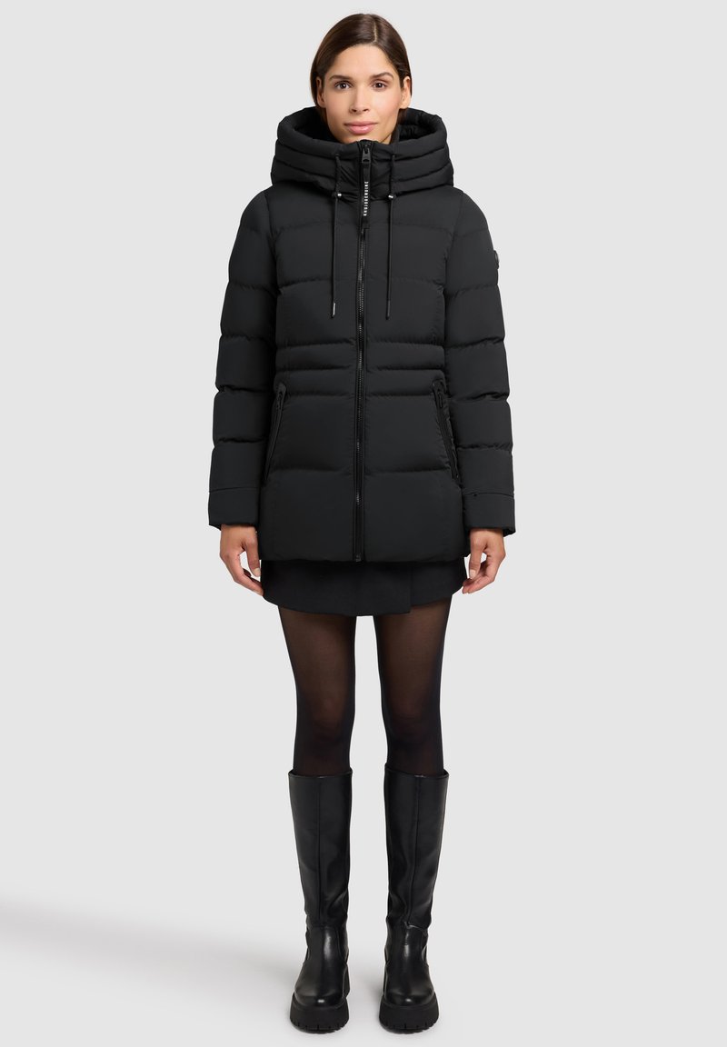 Sort puffjakke med hætte, der har lynlås foran og to sidelommer. Materialet ser blødt ud med et quiltet design og en elegant overflade.