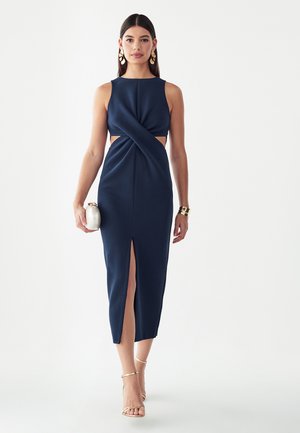 Vrouw draagt een marineblauwe mouwloze midi-jurk met uitsnijdingen, houdt een witte clutch vast en draagt gouden oorbellen, armband en hakken.