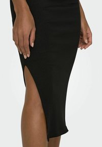 Jupe midi noire à côtes avec une fente latérale, un design ajusté et une texture lisse. Main reposant sur le tissu près du détail de la fente.