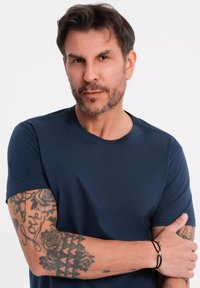 T-shirt blu navy a maniche corte indossato da un uomo con tatuaggi. Presenta un collo tondo e una vestibilità rilassata. Decorato con braccialetti neri al polso.