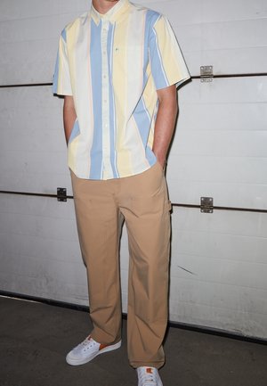 Camisa de manga corta con botones, con rayas azules, amarillas y blancas; pantalones beiges; zapatillas blancas con acentos naranjas.