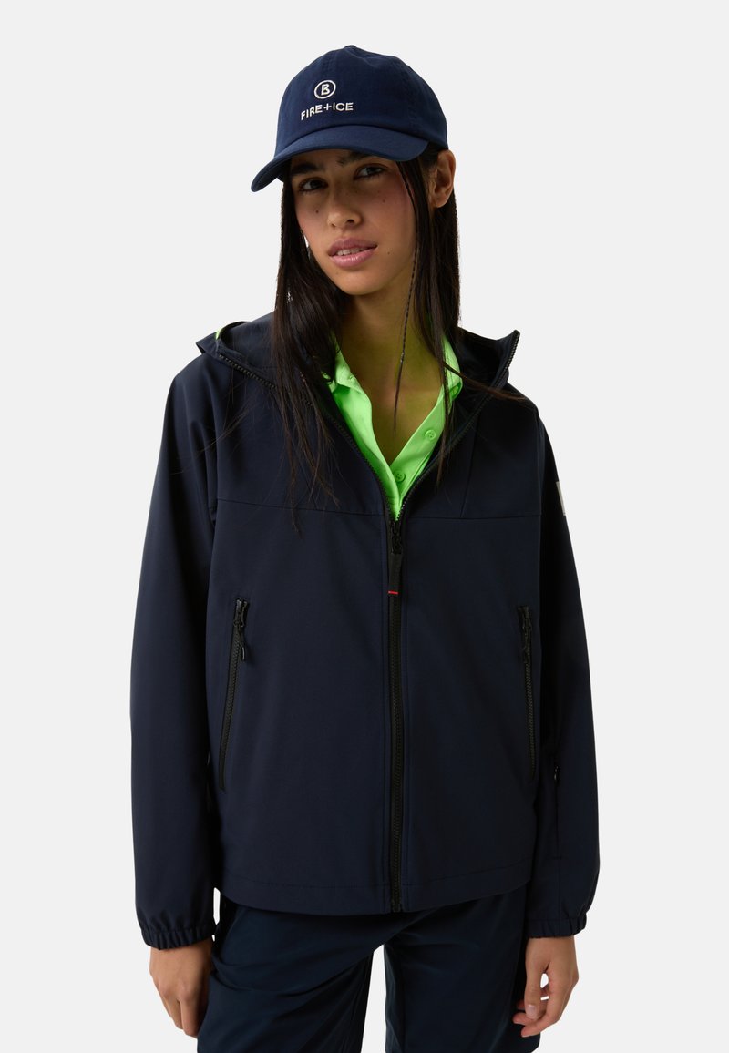 Navy softshell jacket with a zip front, two side pockets, and a hood; paired with a bright green polo and navy cap, creating a layered look.