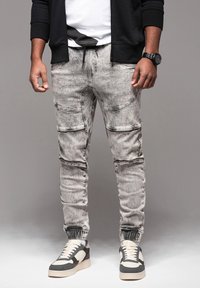 Pantaloni jogger grigi, slim-fit, con polsini elastici, multiple tasche e una texture in denim sbiadito. Abbinati a sneakers in bianco e nero.