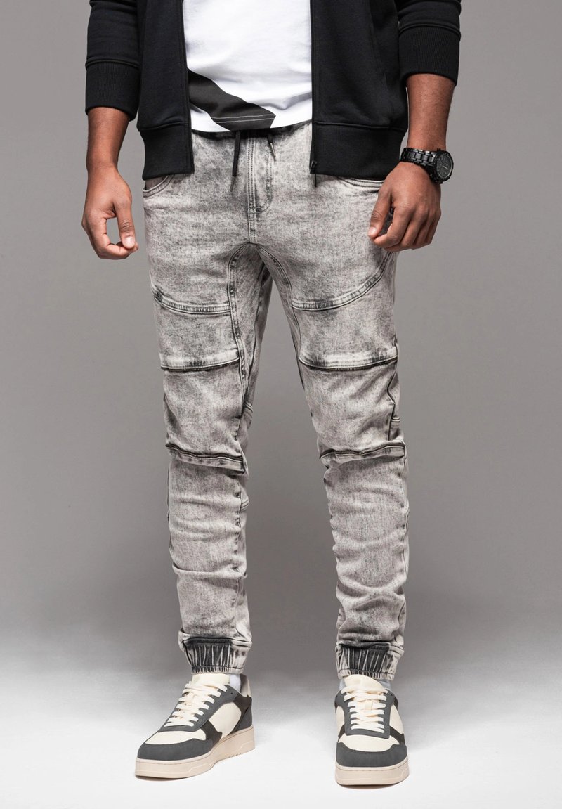 Pantaloni jogger grigi, slim-fit, con polsini elastici, multiple tasche e una texture in denim sbiadito. Abbinati a sneakers in bianco e nero.