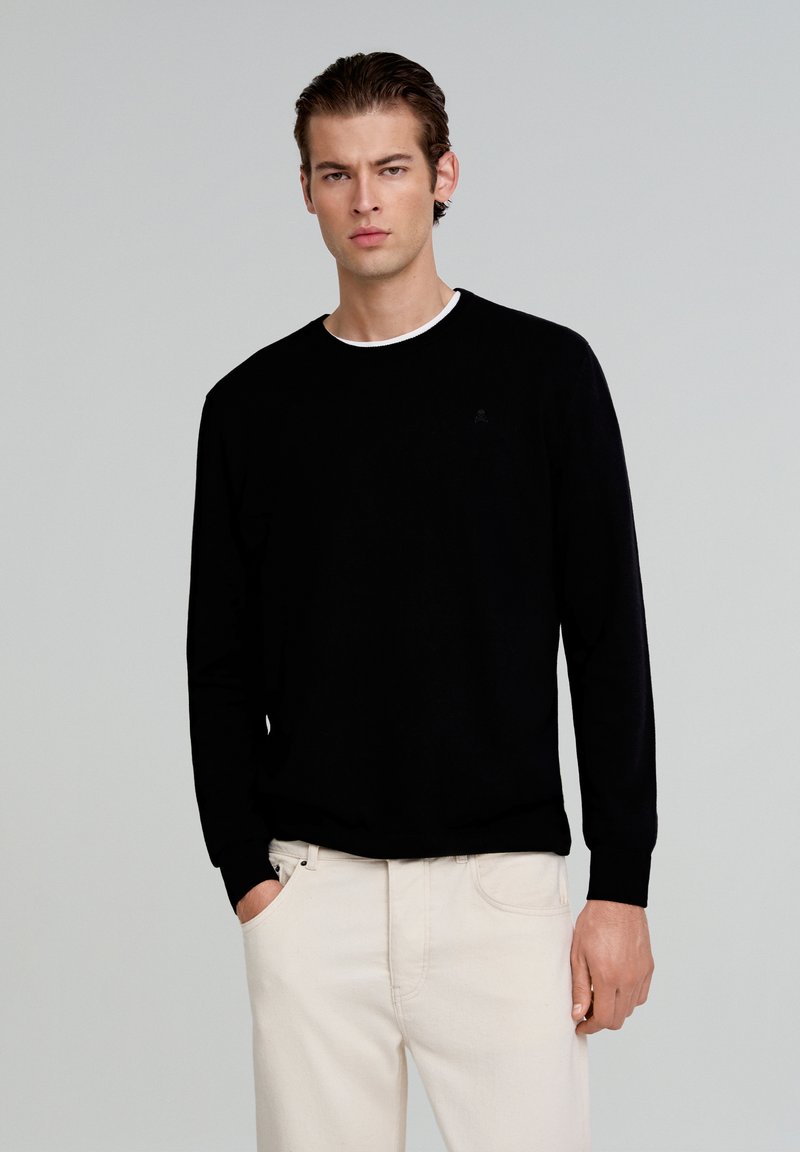 Pull noir à manches longues avec col rond et bord blanc. Texture douce, design ajusté avec des poignets côtelés. Logo discret sur la poitrine.
