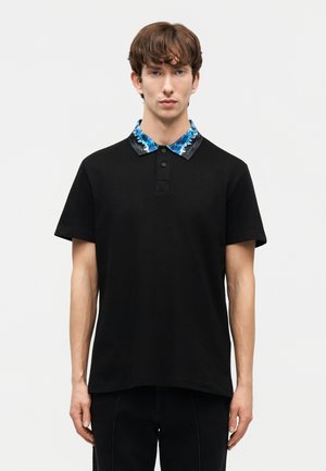 Jonge man met een zwart poloshirt met korte mouwen en een blauw-zwart patroon op de kraag, staande tegen een effen lichte achtergrond.