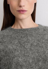 Grijze, fuzzy gebreide trui met een ronde halslijn, voorzien van een zachte textuur. Close-upweergave benadrukt de warmte en details van de stof.