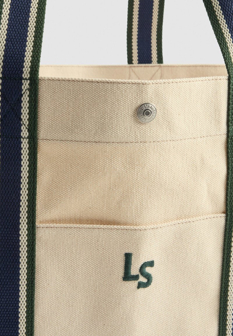 Sac fourre-tout en toile de couleur crème avec des poignées rayées vert foncé et marine. Comprend une petite poche extérieure et un logo brodé "LS" en vert.