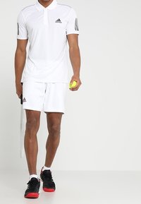 Bílá polokošile Adidas s černými pruhy, spárovaná s odpovídajícími bílými šortkami. Osoba drží tenisový míček a raketu, má na sobě černé boty.