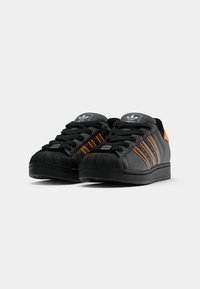 Sneakers pretos com riscas laranja, biqueira em borracha e parte superior em couro texturizado. Apresenta atacadores pretos e logotipo na língua. Design de perfil baixo.