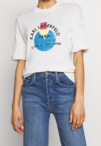 Bílý pletený tričko s barevným grafickým potiskem a textem "Karl Lagerfeld Paris", spárované s modrými džíny. Krátké rukávy a žebrovaný výstřih.