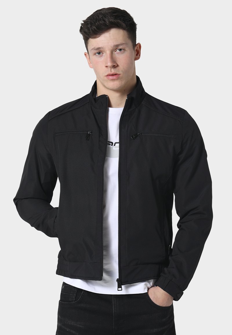 Giovane uomo che indossa una giacca nera con cerniera sopra una maglietta bianca stampata, in piedi con una mano in tasca davanti a uno sfondo semplice.