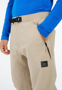 Beige outdoorbroeken met een elastische tailleband en een zwarte gesp, voorzien van zijzakken en een merklogo patch. Gladde textuur, functioneel ontwerp.