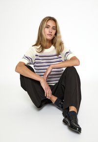 Suéter de punto en crema con rayas moradas, con un escote redondo y mangas cortas. Combinado con pantalones negros holgados y zapatos negros brillantes.