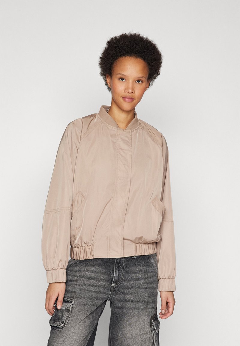 Veste aviateur marron avec fermeture éclair sur le devant, col côtelé, poches latérales, poignets élastiques et ceinture élastique. Porte avec un pantalon cargo gris.