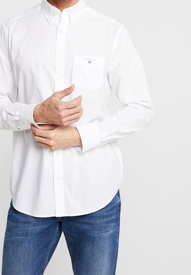 Chemise blanche à boutons avec une poche poitrine, en coton, dotée d'un ourlet arrondi et de manches longues. Portée avec un jean bleu.
