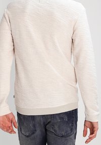 Jasny, fakturowany sweter z okrągłym dekoltem, prążkowanymi mankietami i dołem, w połączeniu z ciemnymi dżinsami. Subtelny wzór dodaje wizualnego zainteresowania.