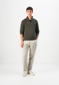 Maglione ribbed verde scuro con colletto a zip, abbinato a pantaloni beige chiaro e sneaker di colore chiaro; design semplice e colori neutrali.