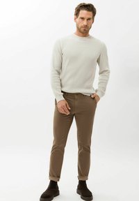 Gebreide crème trui met een gestructureerd patroon, gecombineerd met een strakke bruine broek en donkere enkellaarzen. Model staat zelfverzekerd met de handen in de zakken.