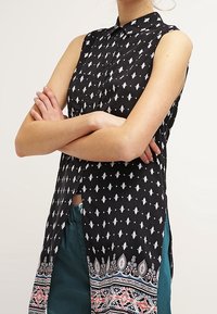 Czarna bluzka bez rękawów z białymi geometrycznymi wzorami, kołnierzykiem na guziki oraz wielokolorowym wzorem paisley na dole. Lekka tkanina.