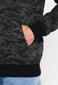 Grå kamuflasje sweatshirt med teksturert stoff, svarte ribbede mansjetter og doble sidelommer med en tettsittende hem.