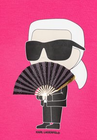 T-shirt graphique représentant un personnage de dessin animé avec des cheveux blancs et des lunettes de soleil, tenant un éventail noir et doré. Fond rose vif, texte noir en dessous.