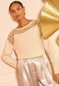 Ženska v bež puloverju z zlatimi bleščicami in sijočih srebrnih hlačah stoji ob svetli steni s zlatim dekorativnim stenovnim umetniškim delom.