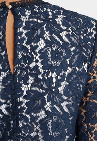 Marinblå blommig spets tyg med nyckelhålsurringning och knäppning på genomskinlig långärmad plagg.